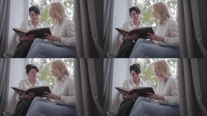 两个成熟的白人妇女坐在窗台上看着照片。资深女性朋友在家分享回忆。快乐微笑的女士们一起在室内度过闲暇时