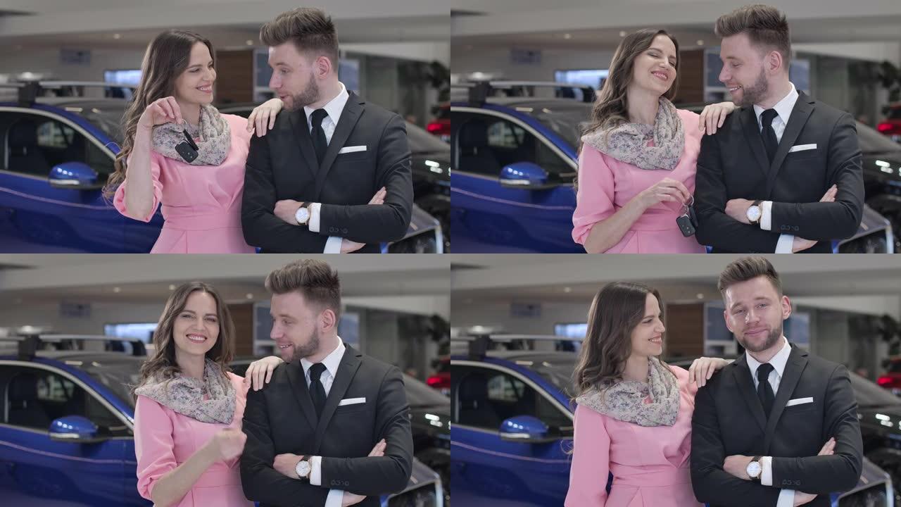 快乐的年轻高加索夫妇在汽车经销店摆姿势的肖像。穿着粉色连衣裙的优雅女人吹牛车钥匙，穿着西装的英俊男人