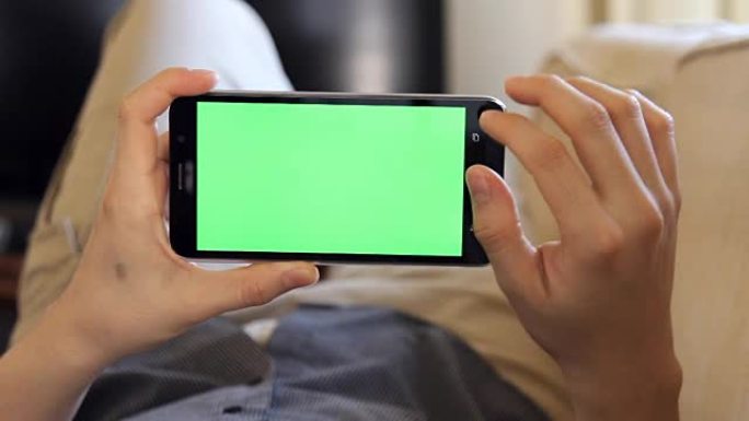 男子躺在沙发上，在风景模式下使用带有绿屏的手机