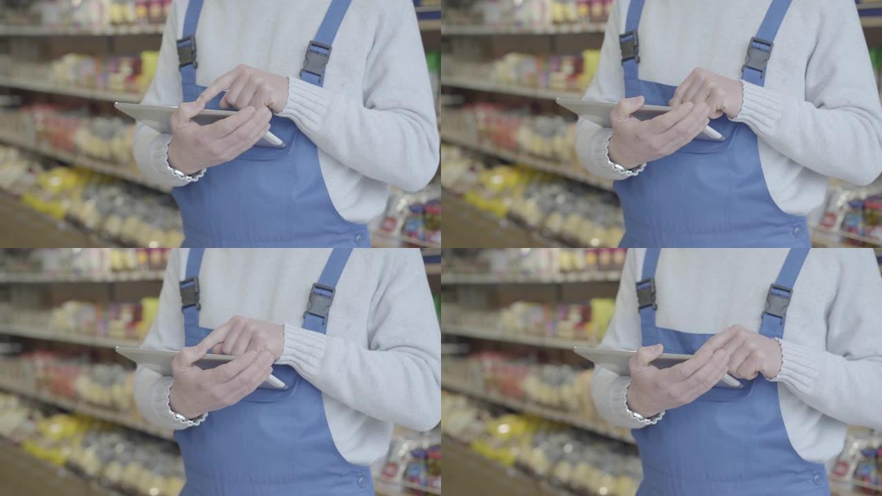 身穿制服的不知名白人男子站在杂货店使用平板电脑。无法识别的人在超市里扫屏检查各种商品。零售。商业，服