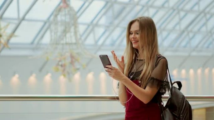 在机场，背着背包的女孩通过耳机和手机在视频上聊天。在智能手机相机上摆姿势自拍，时髦女孩享受广播，在手