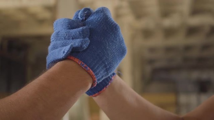 两名戴着蓝色工作手套的白人男性工人的高握手特写。建筑工地强壮的肌肉建设者。