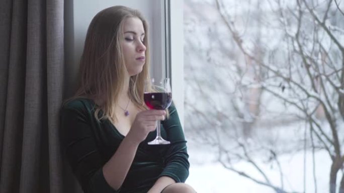 体贴的年轻白人妇女望着窗外，从杯子里喝红酒。孤独美丽的女孩独自在室内度过一天。优雅，生活方式，孤独，