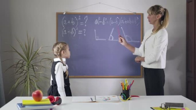 聪明的高加索女人向可爱的小女孩解释数学的肖像。导师指着黑板上写的几何公式说话。教学，教育理念。电影院