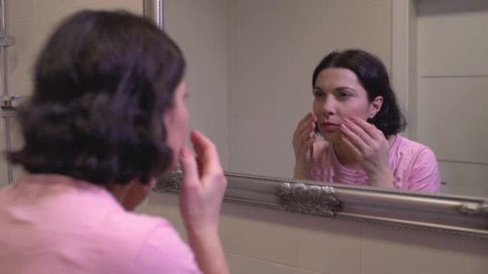 不快乐的中年妇女照镜子，仔细观察脸上的第一条皱纹