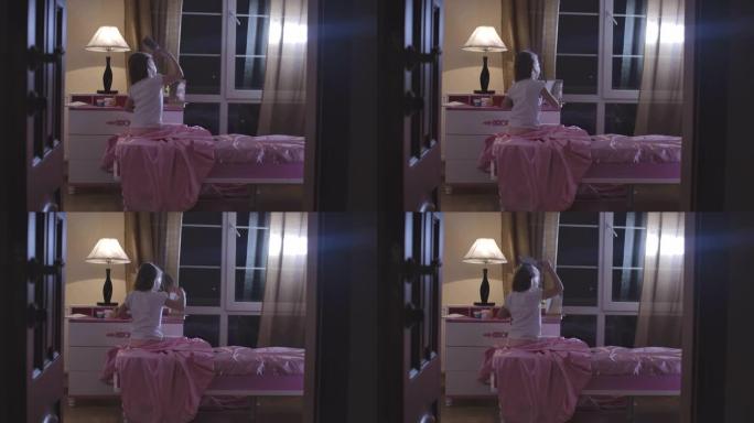 高加索漂亮女孩坐在粉红色的床上梳理头发的后视图。可爱的孩子晚上在她的房间里准备睡觉。青春期，童年，休