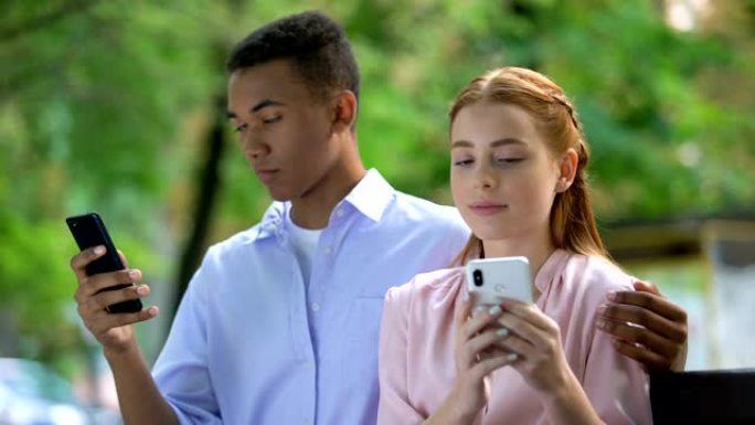 拥抱多种族的青少年夫妇坐在公园的长凳上，聊天智能手机