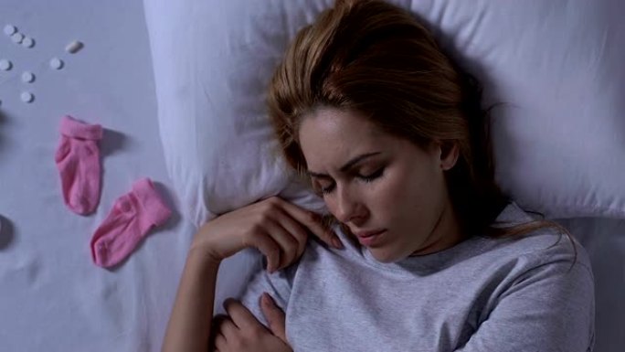 女士躺在床上，靠近粉红色的婴儿袜和流产后自杀的药丸