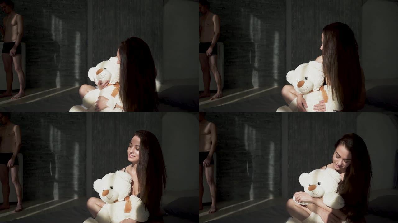 美丽的女孩与黑发在内衣拥抱白色泰迪熊坐在床上。英俊的裸男正站在床边用他的智能手机在手机上输入信息。