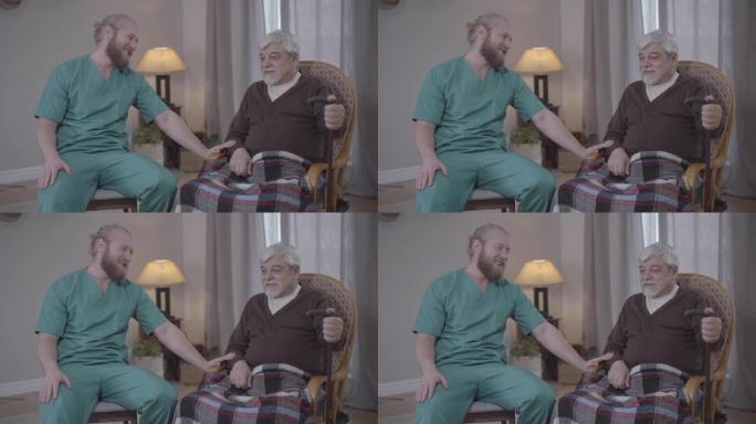 身穿护士服的成年白人男子在疗养院与成熟的男性退休人员聊天。大胡子的年轻人和高级养老金领取者笑着笑。护