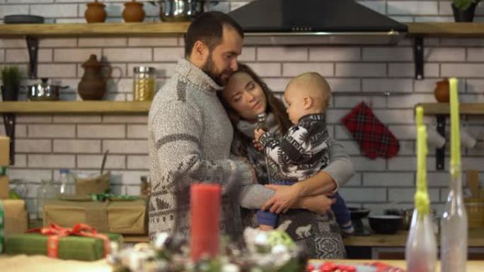 留着胡子的父亲穿着温暖的毛衣，在厨房里和婴儿小儿子在母亲的怀抱里玩耍。