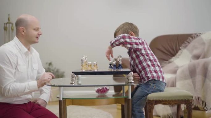 成年高加索人教小儿子下棋的侧视图。可爱的小男孩在家和父亲一起学习。团结、智慧、教育。