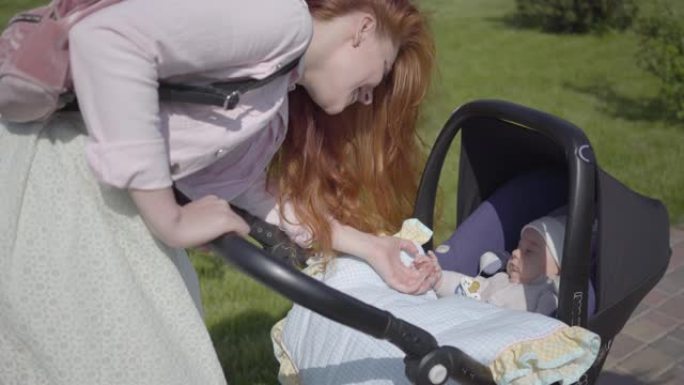 美丽的红发女人弯下腰在公园的婴儿车上。