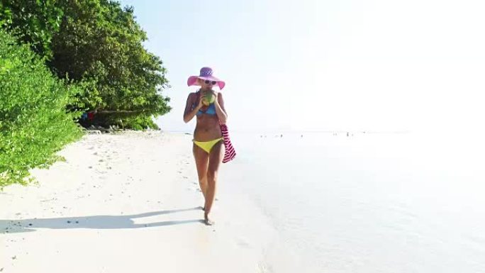 金发女人走在沙滩上，在马尔代夫喝椰子汁