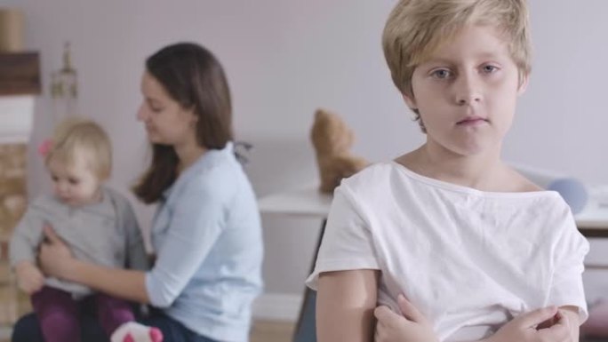 沮丧的高加索男孩的肖像，灰色的眼睛回头看着他的姐姐和母亲在玩耍，转身移开视线。穿着白色t恤的家伙在家