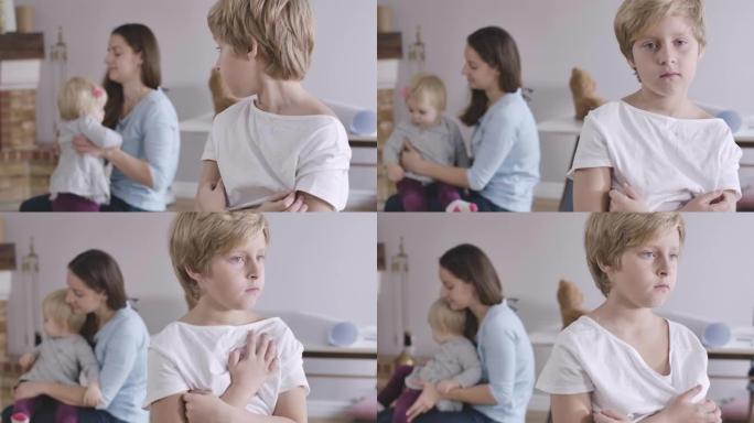 沮丧的高加索男孩的肖像，灰色的眼睛回头看着他的姐姐和母亲在玩耍，转身移开视线。穿着白色t恤的家伙在家