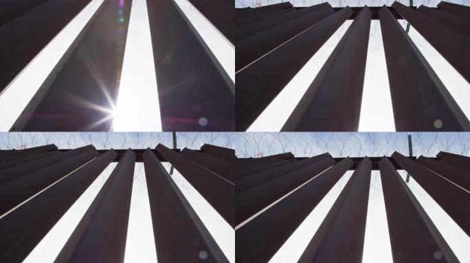 在部分阴天，用剃刀铁丝在墨西哥和美国之间的钢板条边界墙 (在美国一侧) 上窥视太阳的镜头