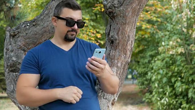 年轻人站在公园的一棵树旁，使用他的智能手机