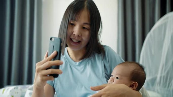 亚洲年轻的母亲和男婴坐在床上，同时通过智能手机进行视频通话