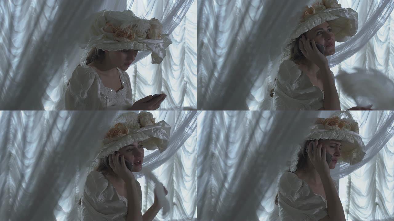 穿着白色复古球裙和白色帽子的年轻漂亮的傻笑女人站在透明窗帘的背景上，挥舞着风扇在手机上说话