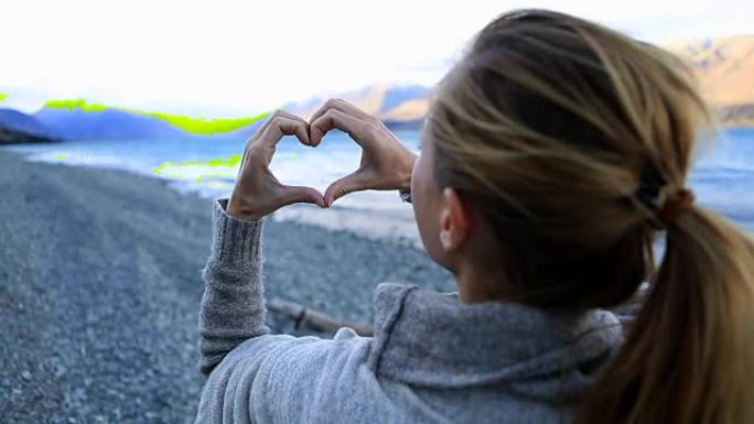 湖岸旁的年轻女子在山湖景观上制作心形手指框