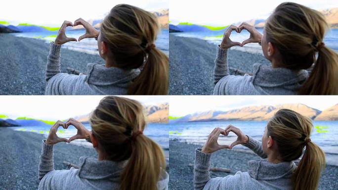 湖岸旁的年轻女子在山湖景观上制作心形手指框