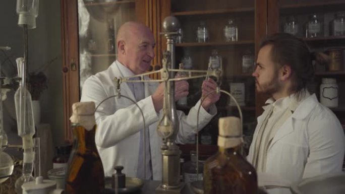 快乐的白人老人向年轻人展示老式试管和烧瓶并交谈。穿着白袍的药剂师在古代药房工作。19世纪，复古药店。