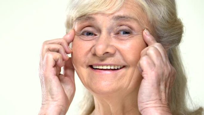 微笑的老妇人拉伸眼部皱纹，面部轮廓抗衰老按摩