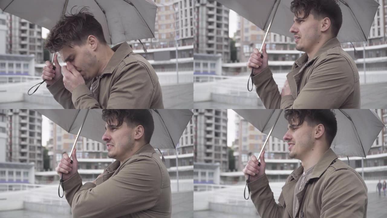 年轻患病的白人男子的特写镜头，他拿着雨伞站在户外，打喷嚏和咳嗽。英俊的黑发男人在城市街道上等待见面的
