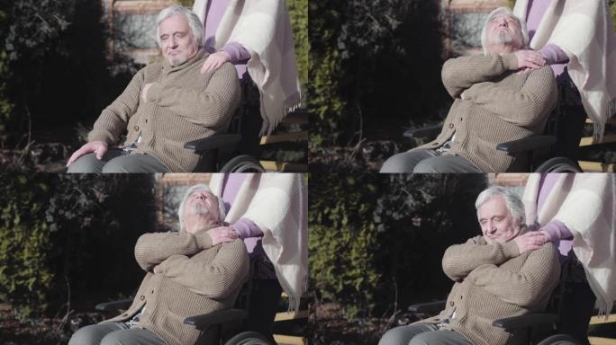 疲惫的白人残疾老人的肖像坐在户外的轮椅上，牵着无法辨认的护士的手，握住他的肩膀。养老院的概念，无效招
