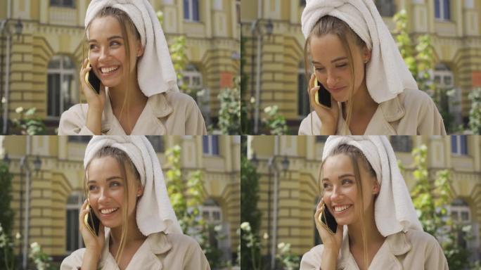 穿着浴袍的漂亮微笑女孩的肖像，头上有毛巾在街上用手机说话。
