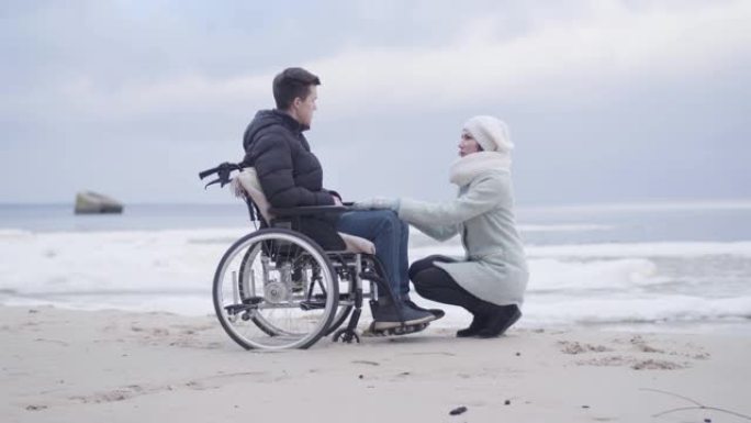 年轻女子坐在轮椅上的男孩面前聊天。母亲或无效的温柔支持户外海岸或河岸上的残疾年轻人。快乐，生活方式，