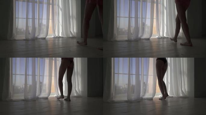 性感的女人的腿走到地板到天花板的窗户和脱下睡衣。