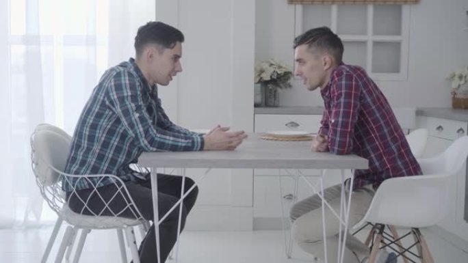 两个成年的高加索双胞胎兄弟坐在桌旁，彼此交谈。友好的兄弟姐妹在家里共度一天。家庭，友谊，友谊。