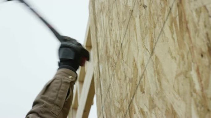 一名四十多岁的拉丁裔男子在冬天阴云密布的天空下，用锤子将一块刨花板固定在建筑工地上。