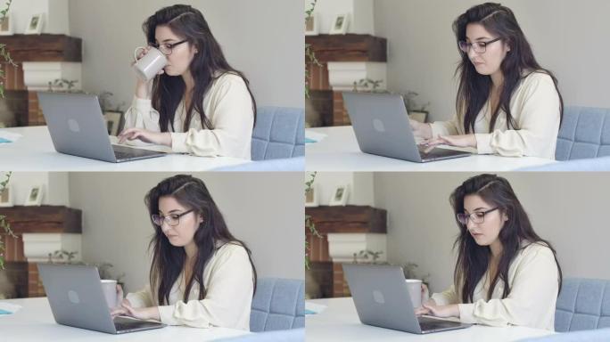 年轻聪明的白人妇女在眼镜上喝茶或咖啡，在笔记本电脑上打字。自信的聪明女孩，长长的黑发和棕色的眼睛在家