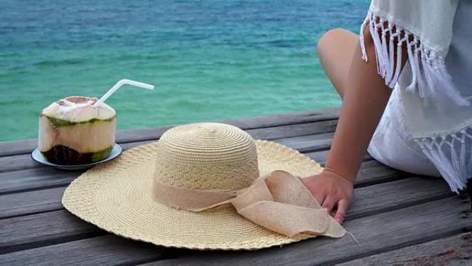 女人戴着一顶海帽，她很开心地坐着，椰子在木桥上喝酒，看着海滩海边，云和蓝天是无尽的背景，复制空间。