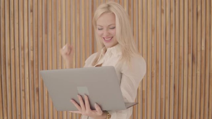 美丽的白人妇女，金发和灰色的眼睛拿着笔记本电脑，做出胜利的手势