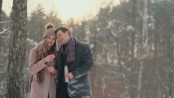 幸福的夫妇拿着热茶杯在冬天的风景。热恋中的年轻夫妇放寒假，站在一棵树旁，喝着热腾腾的茶