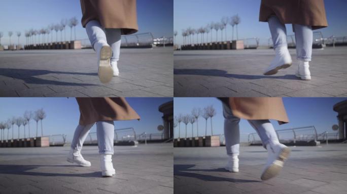 摄像机跟随穿着牛仔裤和白色运动鞋的女性腿走在城市广场上。年轻女子在阳光明媚的日子在户外漫步。生活方式