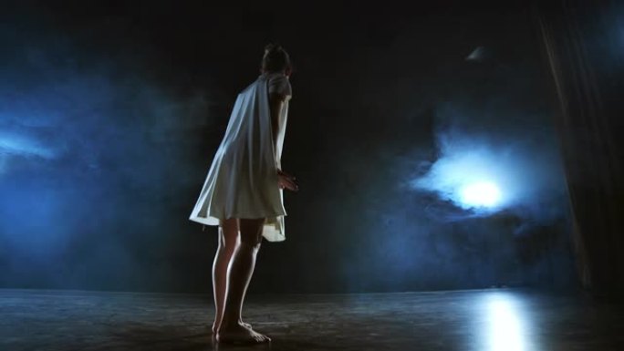 一个穿着白色连衣裙的女人在舞台上跳舞着现代芭蕾舞的戏剧性舞蹈。芭蕾舞演员在烟雾中移动塑料表演现代舞蹈