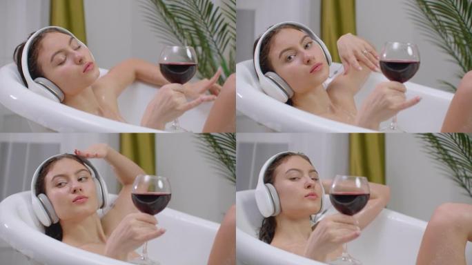 放松的年轻女子听着音乐在一个泡泡巴。一个放松的年轻女子听音乐耳机在一个泡沫浴和喝红酒从一个杯子。