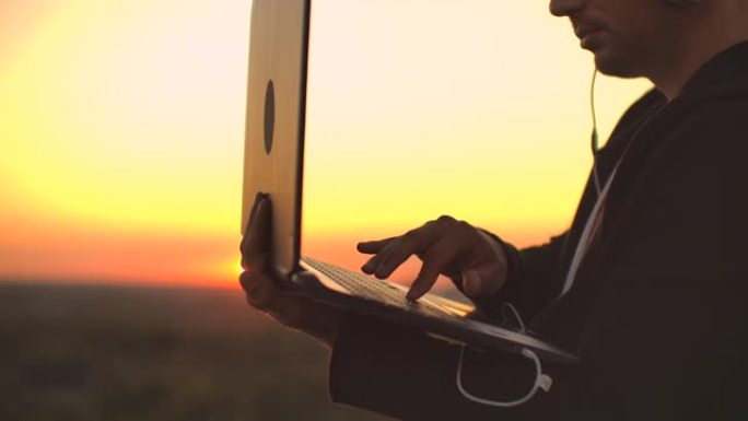 日落时分站在屋顶上拿着笔记本电脑和啤酒。一名身穿帽衫的男子在高处欣赏着美丽的城市风景