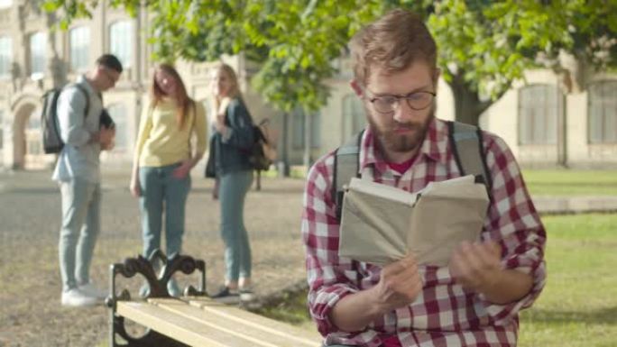 害羞的白人大学生戴眼镜，遭受同伴的欺凌。年轻的大胡子男人坐在长凳上的肖像，书当模糊的人在背景上大笑。