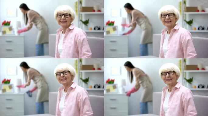 微笑的老年妇女看着相机，管家在房间里擦灰尘，打扫卫生