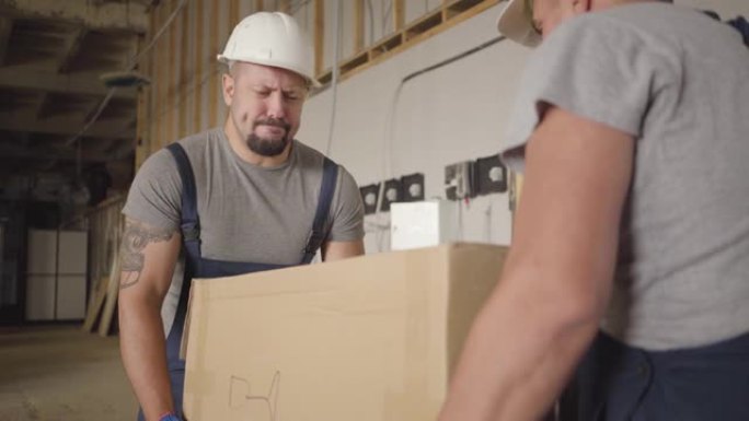 两名强壮的高加索工人拿起沉重的纸板箱，在施工现场搬运。穿着防护头盔和制服的成年男性建筑商在室内工作。