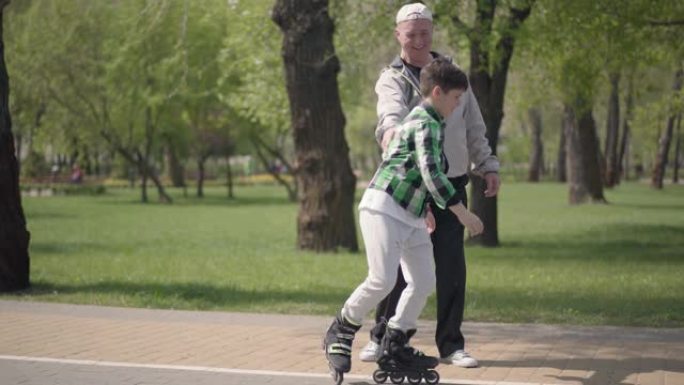 可爱的男孩穿着方格衬衫在公园里溜旱冰，他的祖父抓住他并拥抱他。