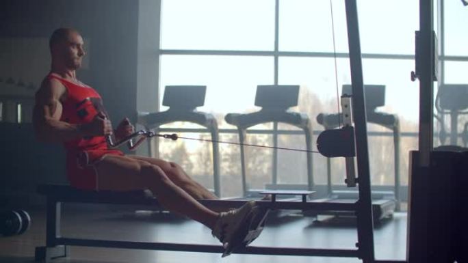 一名男子坐在模拟器中，在健身房大窗户的背景下，对背部肌肉进行锻炼，将体重拉到腹部。有实力的男运动员划