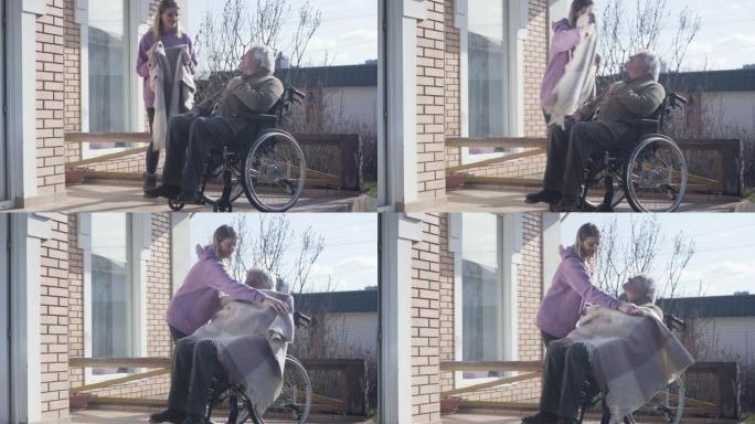 戴眼镜的白人妇女为坐在轮椅上的残疾老人带毯。护士照顾享受阳光明媚的秋日的老退休人员。帮助，帮助，支持