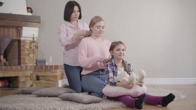 母亲给大女儿编织辫子，而给小女孩编织辫子。快乐的高加索家庭在室内一起休息。关心、支持、生活方式。